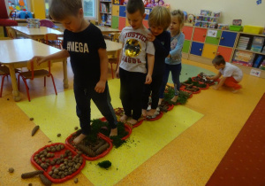 Na podłodze ułożone są tacki z różnorodnym materiałem przyrodniczym po których przechodzi czworo dzieci bosymi stopami. W tle chłopiec przykuca przy tacce, układa materiał przyrodniczy.